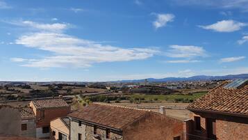 Paisaje rural con edificios de tejados terracota, una cadena montañosa distante y un cielo azul despejado.