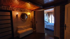 A cozy room with a wooden ceiling, a white sofa, a round mirror, and a door leading to a bedroom with a bed and curtains.