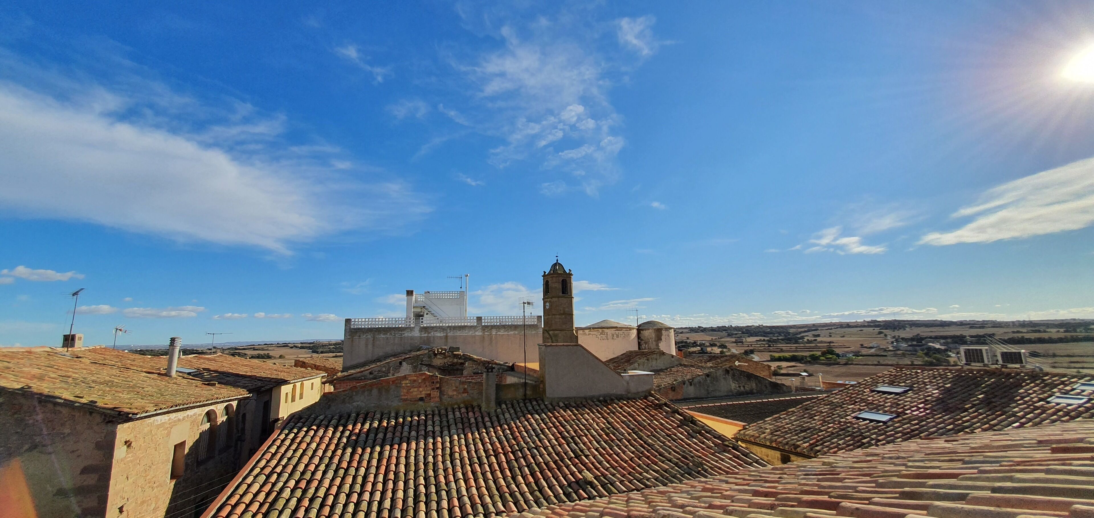Una vista panoramica di una città storica con tetti in tegole di terracotta, un’imponente torre campanaria e un cielo azzurro intenso.