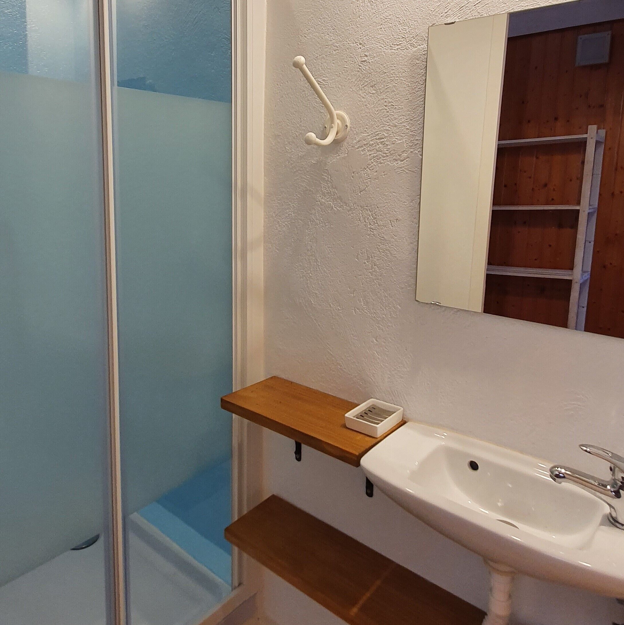 Un baño con un lavamanos blanco, un estante de madera, un espejo y una zona de ducha.