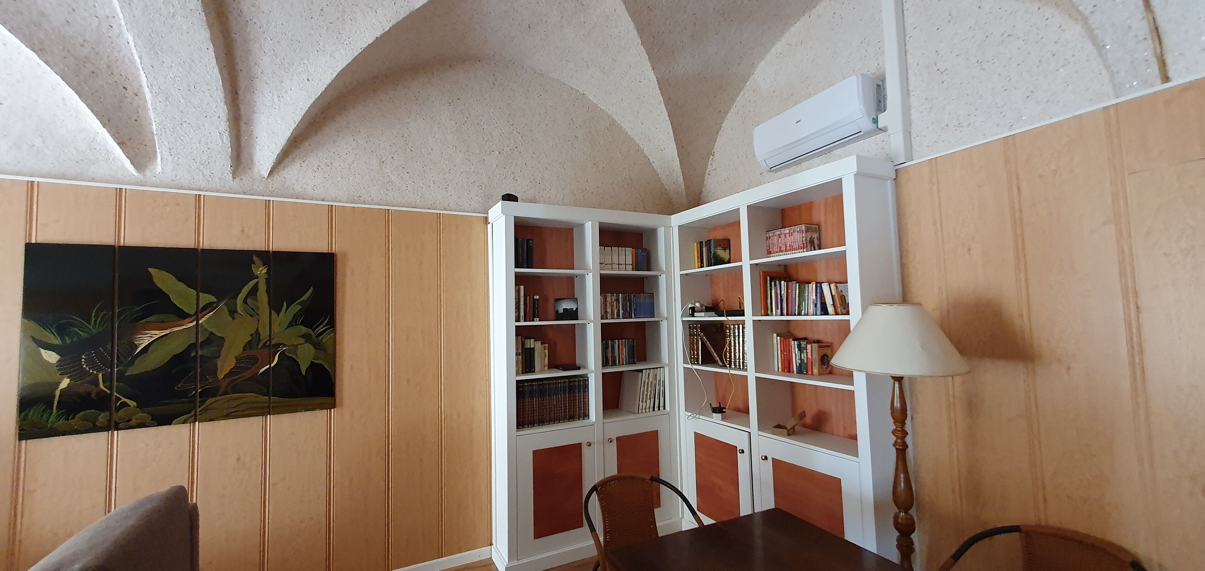 A room with wooden panel walls, a white bookshelf, a table with chairs, and a ceiling with arched designs.