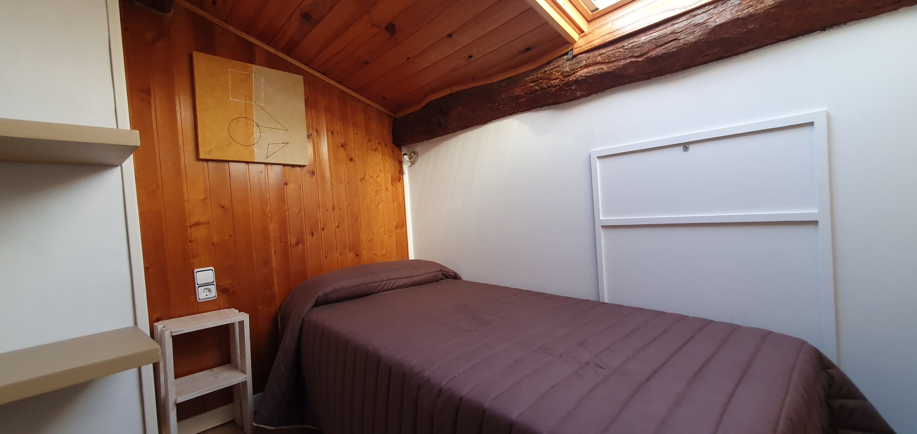 A small, cozy room with wooden walls, a single bed with a brown bedspread, a white shelf, and a white door.