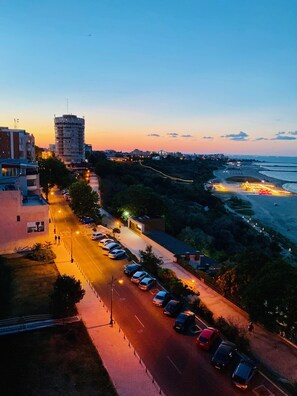 Un paysage urbain côtier au crépuscule, avec une route bordée de voitures garées, des bâtiments et une plage en arrière-plan.