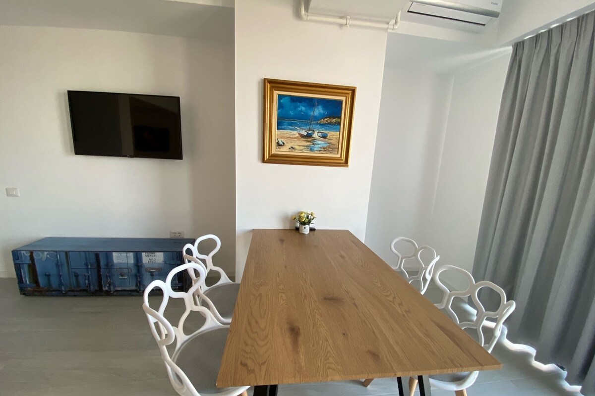 A room with a wooden table, white chairs, a painting, a TV, and a blue cabinet.