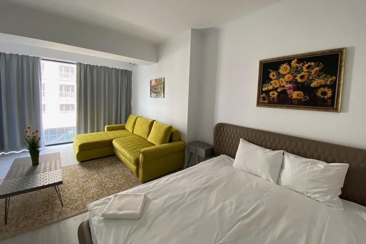 A hotel room with a bed, a yellow sofa, a small table with a vase, and a painting on the wall.