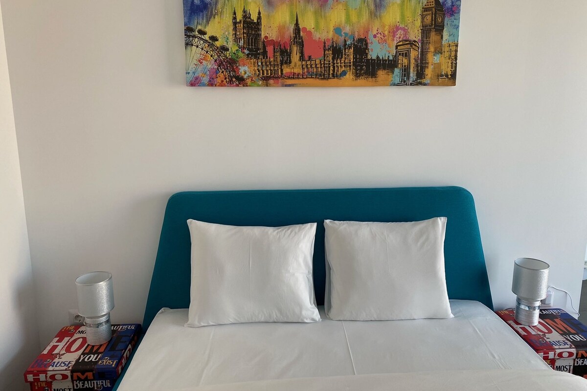A hotel room with a teal headboard, white bedding, and a colorful cityscape painting above the bed.
