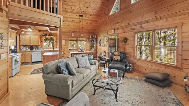 A cozy cabin interior with a living room, kitchen, and dining area, featuring wooden walls and large windows. - Timber Lodge - Private, Serene Cabin with Game Room for Kids (Todd)