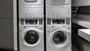 Two stacked front-loading washing machines in a laundry room with visible control panels and a ceiling with exposed pipes and lighting fixtures. - Downtown Retreat: Stylish Boston Studio (Boston)