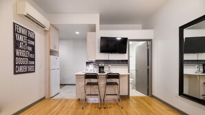 A modern kitchen with a central island, two bar stools, a flat-screen TV, and a wall-mounted air conditioner. - Downtown Retreat: Stylish Boston Studio (Boston)