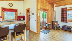 A cozy cabin interior with a wooden ceiling, a kitchen with a red window blind, a dining area with two chairs, and a fireplace with logs. - 2 bedroom accommodation in Torlundy, near Fort William (Torlundy, near Fort William)