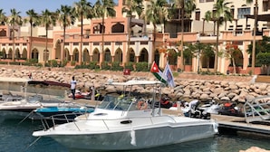 A marina with boats docked, a rocky shoreline, and a building with arches in the background. - Bait Alaqaba Resort and Dive Center (Aqaba)