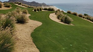Um campo de golfe com um caminho de areia que leva ao oceano, cercado por relva verde e arbustos.
