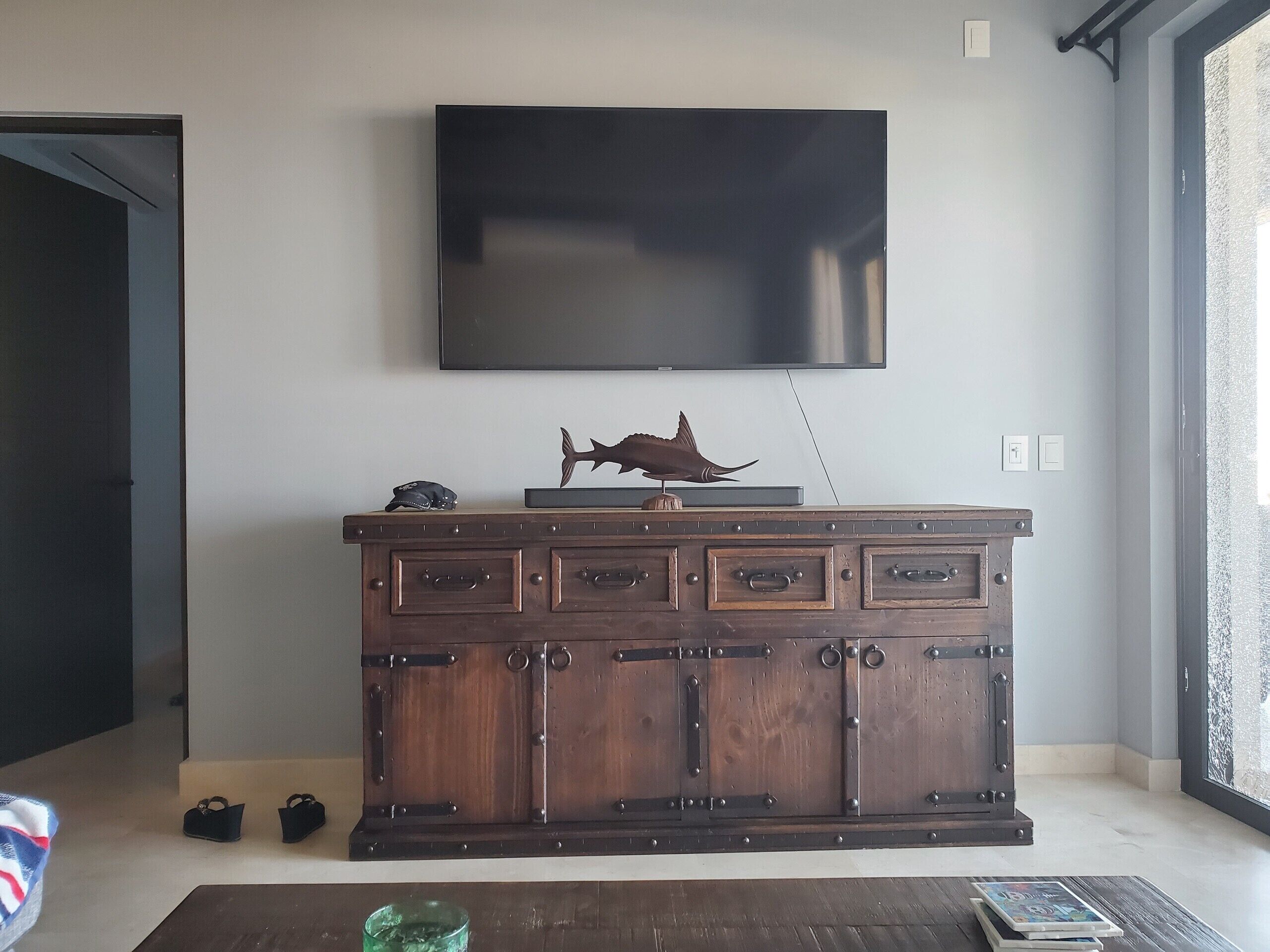 Un salon avec un téléviseur à écran plat fixé au mur, un meuble en bois doté de plusieurs tiroirs et une paire de chaussures posées sur le sol.
