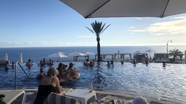 Uma área de piscina com vários guarda-sóis e espreguiçadeiras, com vista para o mar azul e límpido.