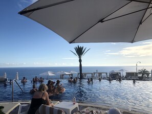Zona de piscina con múltiples sombrillas y sillones de descanso, con vista al mar azul y despejado.