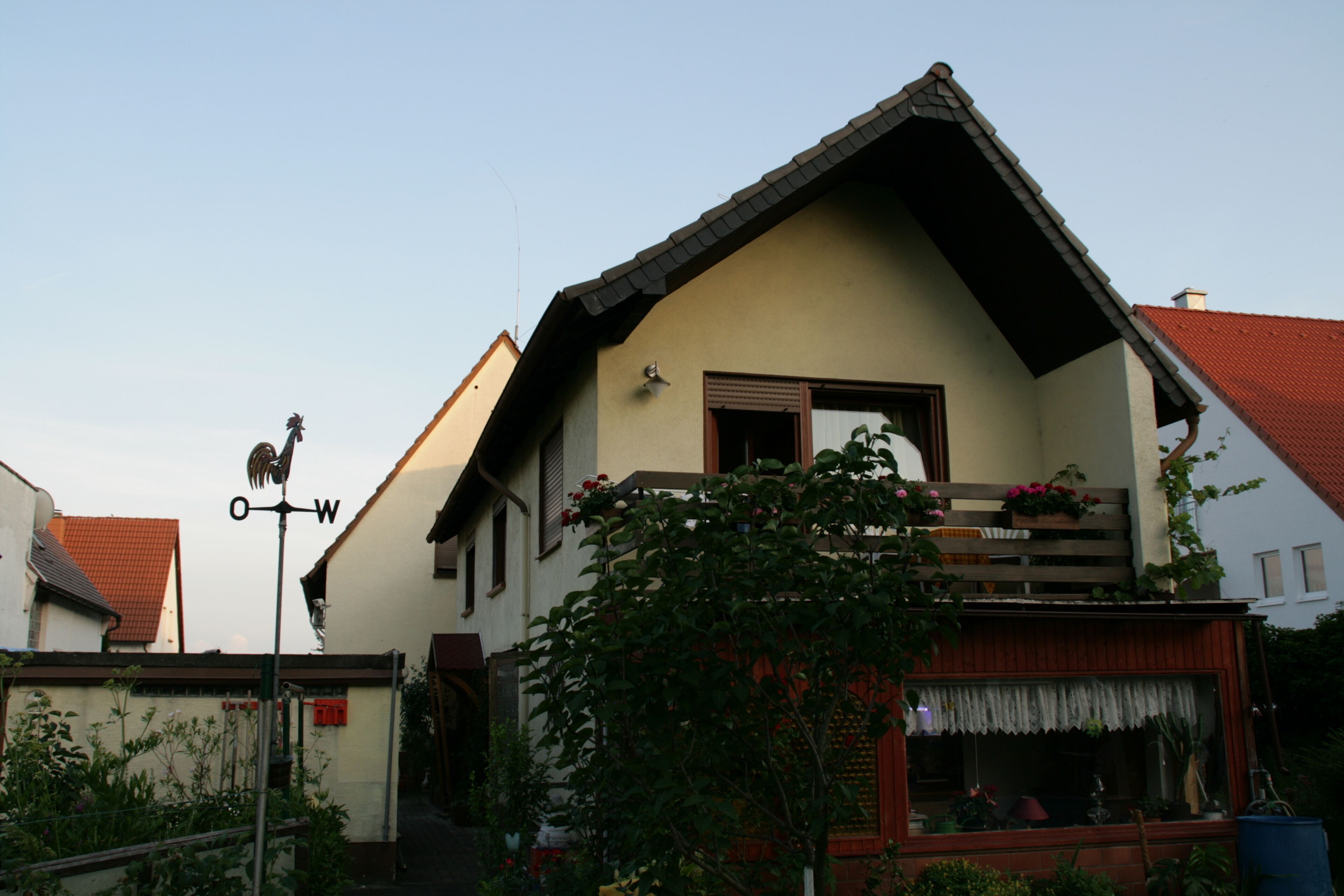A two-story house with a sloped roof, a balcony with flowers, and a visible weather vane.