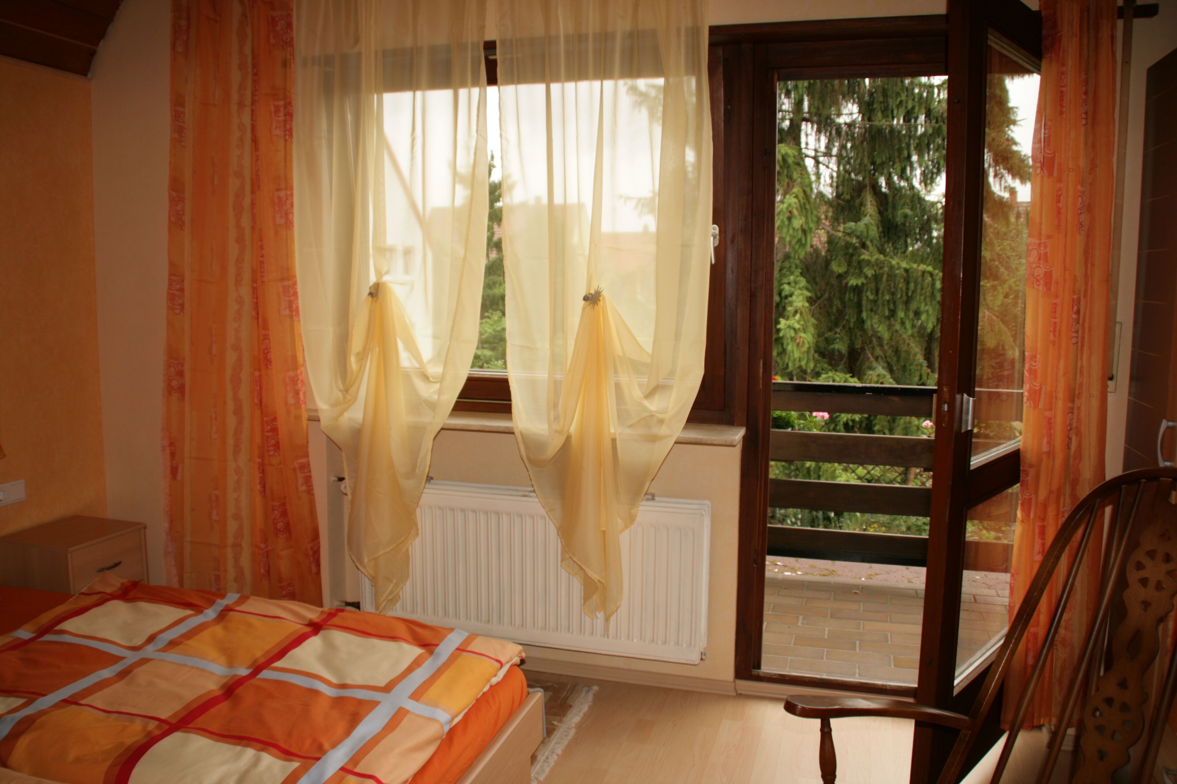 Une chambre à coucher comprenant un lit, une chaise, une fenêtre avec des rideaux, un radiateur et une vue sur la verdure à l’extérieur.