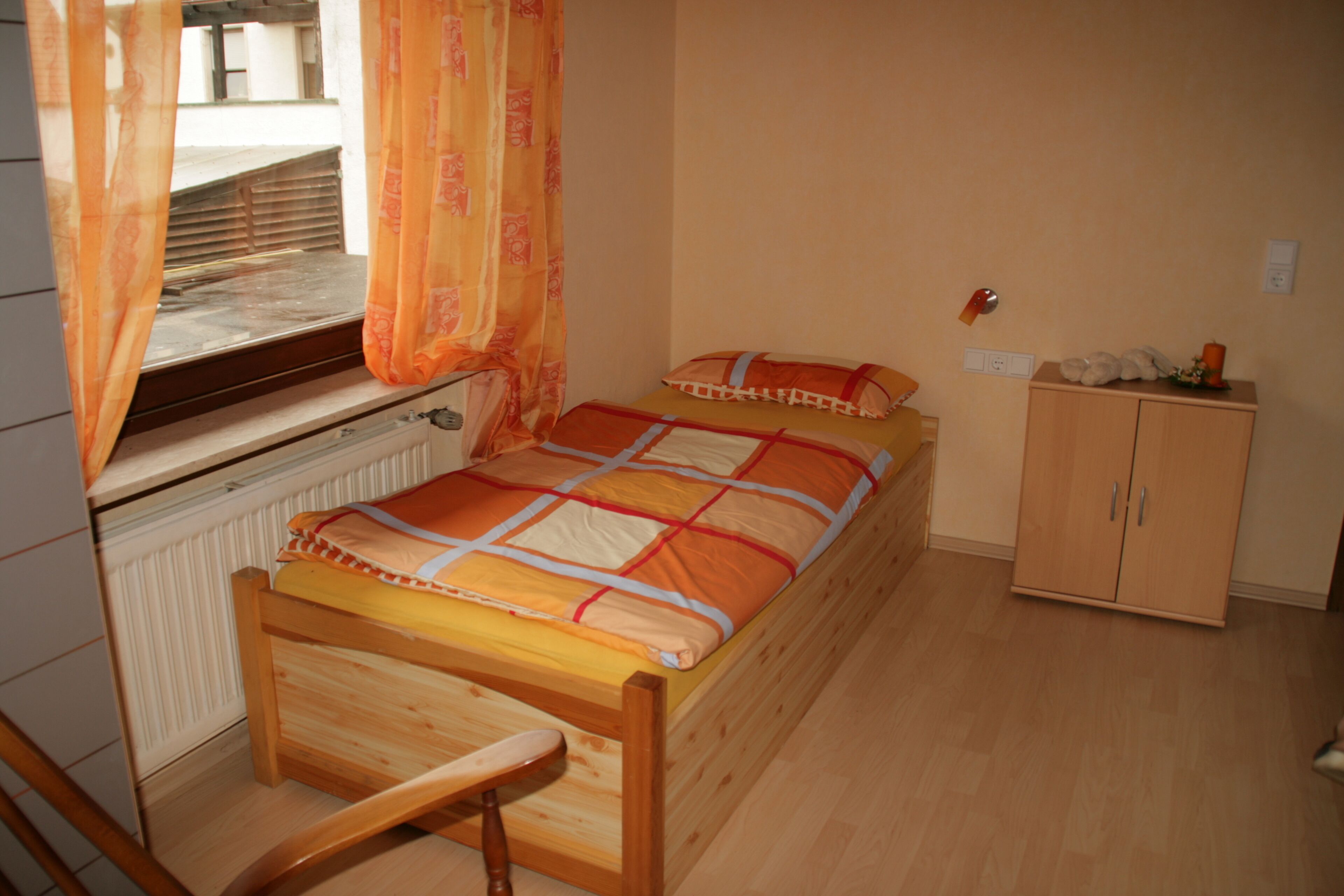 Une chambre avec un lit en bois, une fenêtre avec des rideaux, un radiateur, une armoire en bois et une chaise.