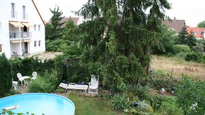Un patio trasero residencial con piscina, un porche amoblado y una diversidad de plantas y árboles.