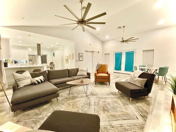 A modern living room with a sectional sofa, a coffee table, and a small armchair. There are two ceiling fans, a dining area with a table and chairs, and a kitchen in the background. - Bayshore Beach House - Walk to Nokomis Beach!  Close to Siesta Key, Venice Beach (Nokomis)