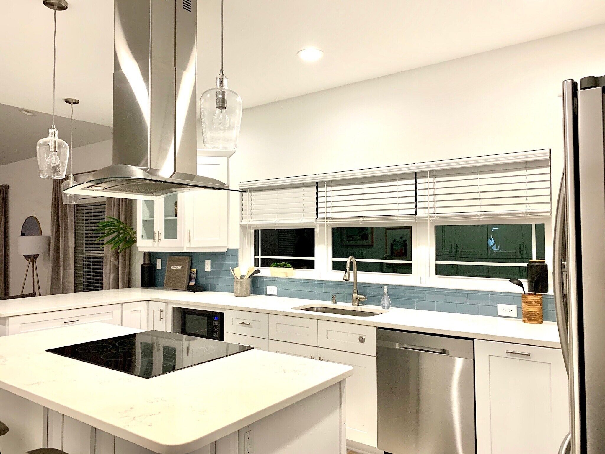 A modern kitchen with white cabinetry, a stainless steel dishwasher, a built-in oven, and a window with blinds.
