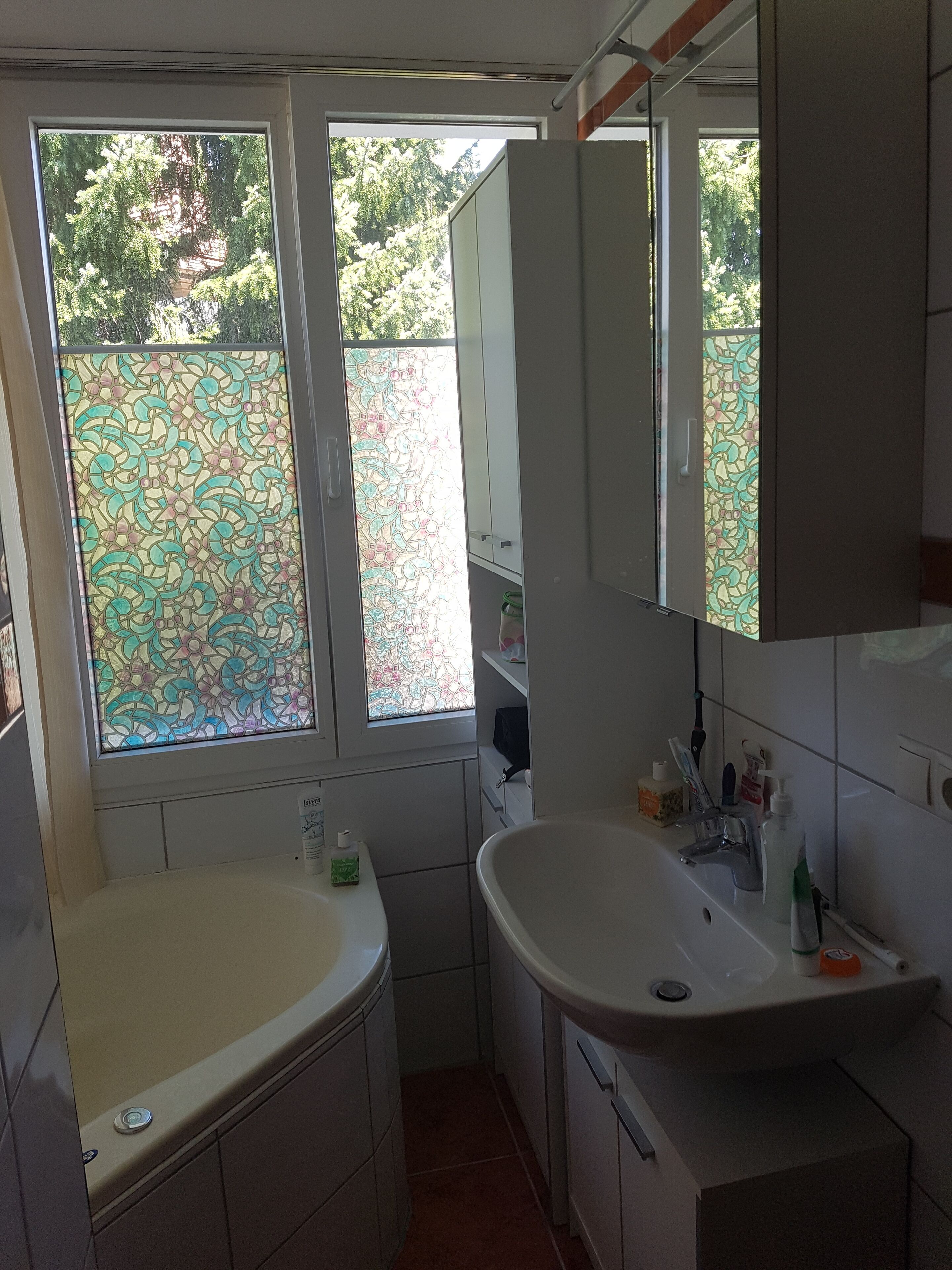 Une salle de bain avec une baignoire, un lavabo et un grand miroir. Il y a des fenêtres avec des vitres décoratives.
