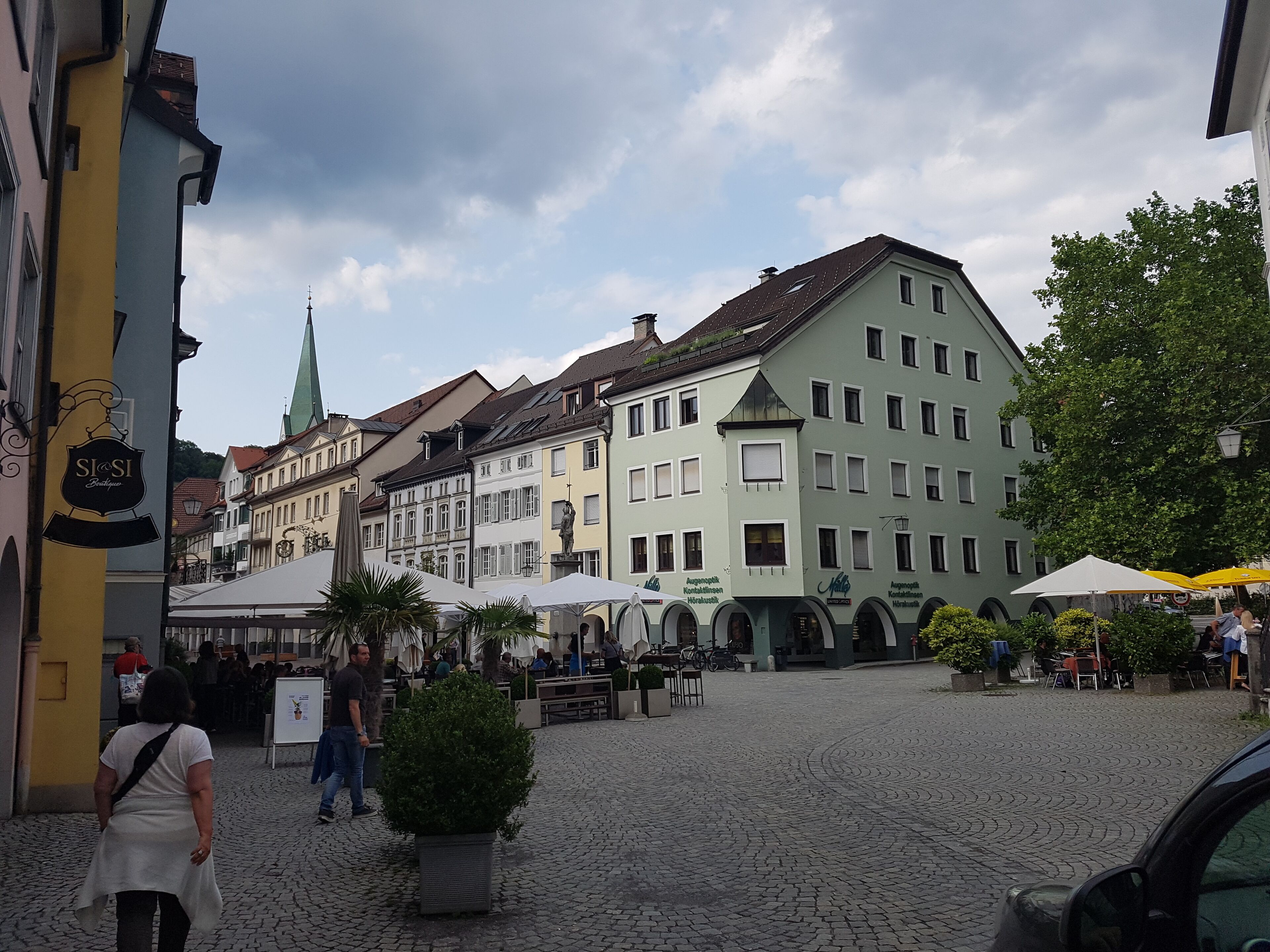 Ein Kopfsteinpflasterplatz mit historischen Gebäuden, Außenbestuhlung und einer Statue.