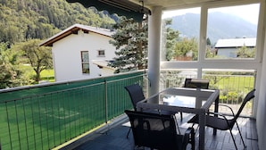 Ein Balkon mit Tisch und Stühlen, einem grünen Zaun und Blick auf ein Haus und Berge.