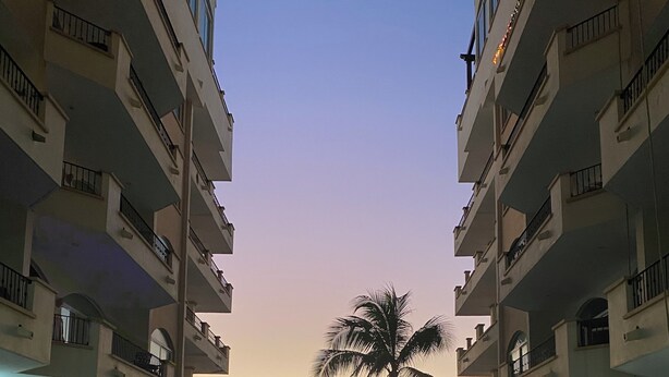 A resort pool with a palm tree, surrounded by multi-story buildings with balconies and outdoor seating. - Resorto Condominio frente al mar directamente en el océano - Vista marina (Marina Mazatlán)