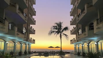A resort pool with a palm tree, surrounded by multi-story buildings with balconies and outdoor seating.