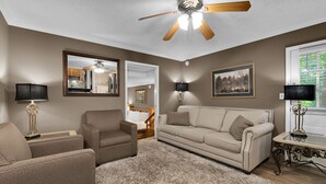 A living room with a beige sofa, a brown armchair, a ceiling fan, and a large window with blinds. - Eureka Uptown Garden Cottage (Eureka Springs)