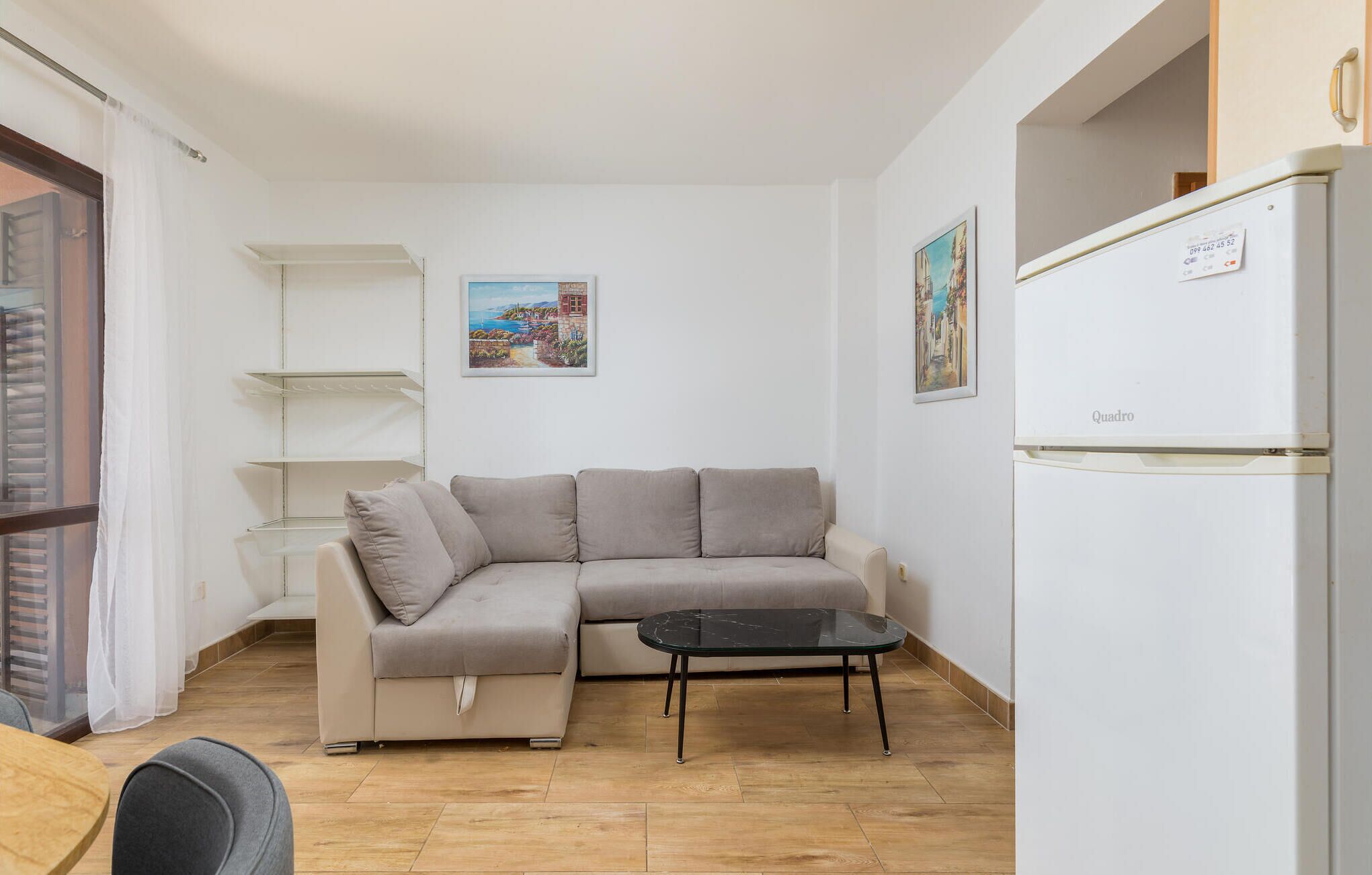 Ein kleines, modernes Wohnzimmer mit einem beigefarbenen Ecksofa, einem Glastisch und einem weißen Kühlschrank.