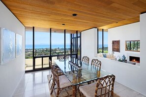 Enjoy meals in this dining room with a glass-top table and wicker chairs, surrounded by a high ceiling and large windows offering a view of the landscape.
