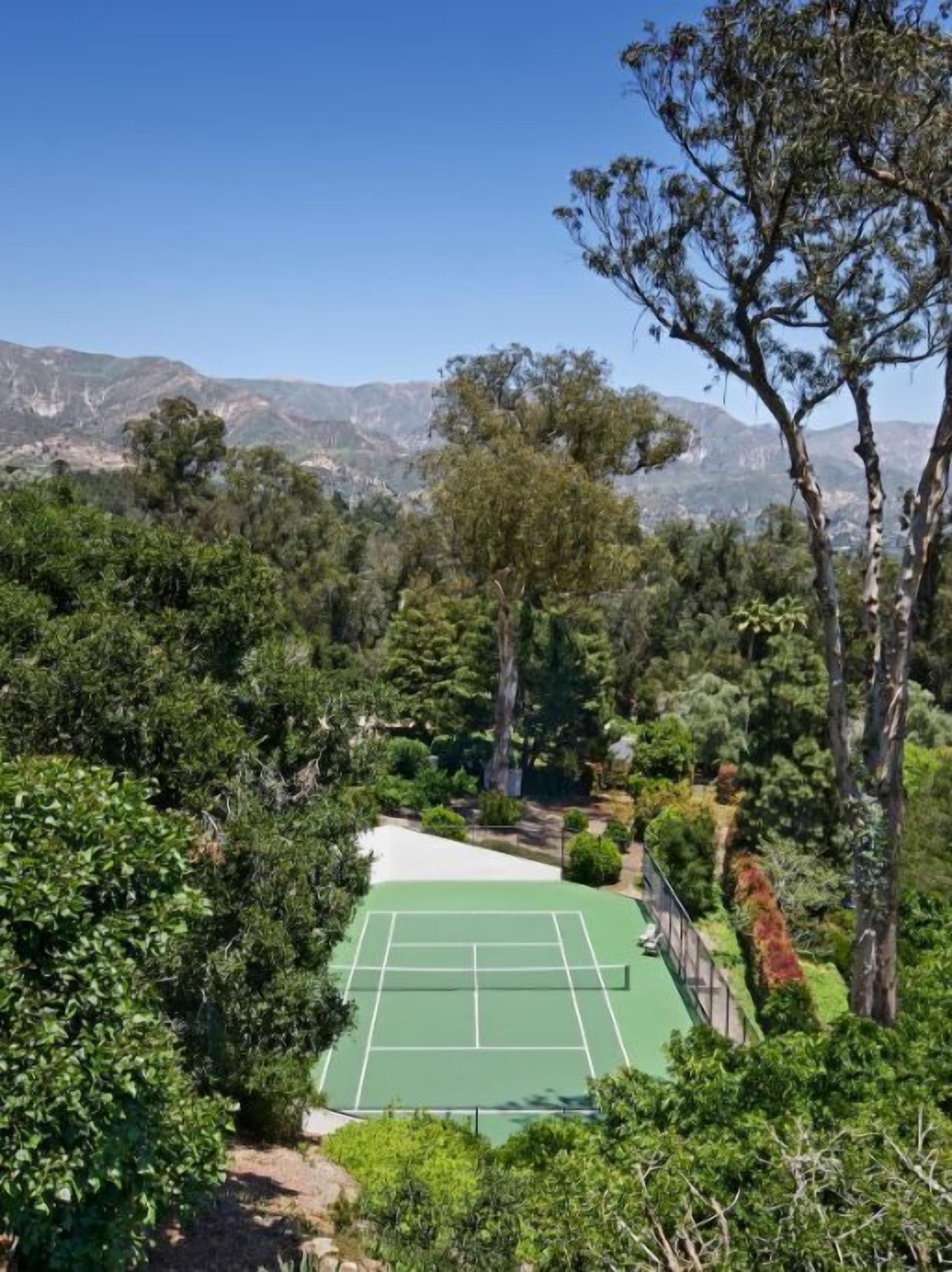 A tennis court surrounded by dense greenery and mountains in the background.