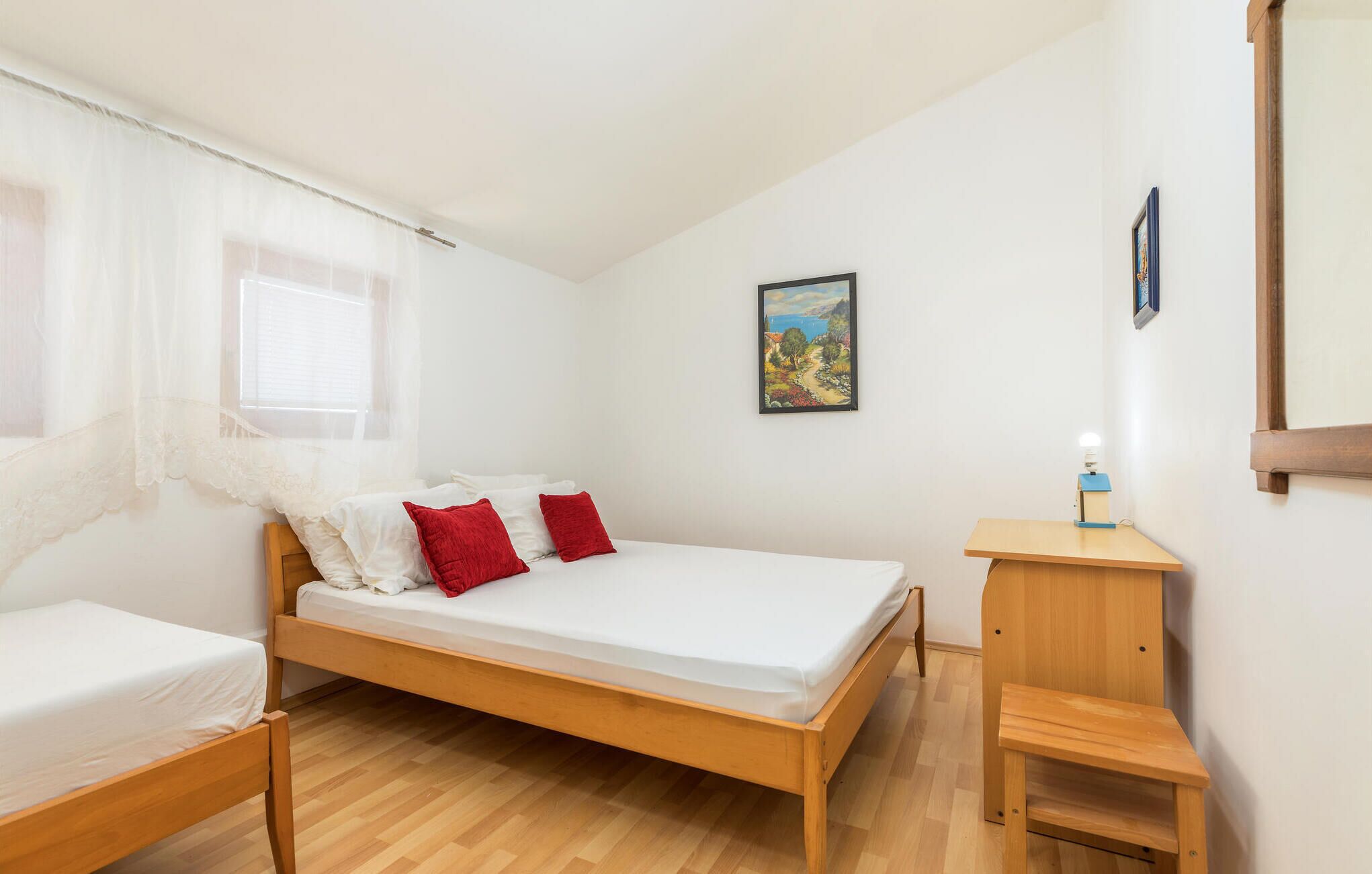 Une chambre avec un lit simple, un cadre de lit en bois, une petite table de chevet, une commode, une fenêtre avec des rideaux légers et un tableau au mur.