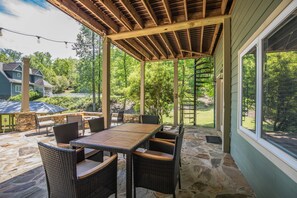 An outdoor patio with a wooden table and wicker chairs, stone flooring, and a view of a green landscape. - Secluded Lake Home (Dadeville)