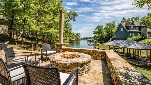 A stone fire pit surrounded by wicker chairs, a gazebo, and a view of a lake with a house in the background. - Secluded Lake Home (Dadeville)