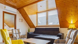 Een knus loft-achtig vertrek met een schuine kap, een bank, twee fauteuils, een kleine tafel en een raam met zonwering.