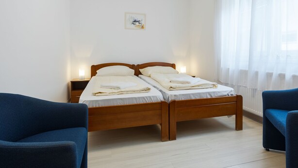 Een slaapkamer met twee houten bedden, een nachtkastje, een blauwe fauteuil en een raam met dunne gordijnen.