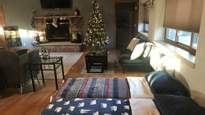 Ein gemütliches Wohnzimmer mit Kamin, Weihnachtsbaum, einem Bett mit gemusterten Bettbezügen und einem Fenster mit Jalousien.