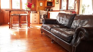 A cozy living room with a leather sofa, wooden flooring, a kitchen area with a stove and cabinets, and large windows. - Welcome Home Cottage, Manapouri, Fiordland (Manapouri)