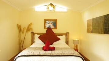 Una cama bien hecha, con una almohada roja y toallas enrolladas, una lámpara de noche pequeña y un cuadro en la pared.