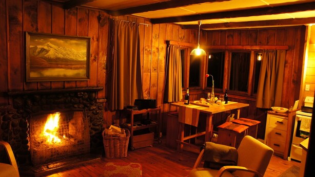 A cozy room with a stone fireplace, wooden walls, a dining table, and a kitchen area. - Welcome Home Cottage, Manapouri, Fiordland (Manapouri)