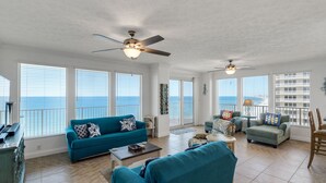 Ein geräumiges Wohnzimmer mit einer großen Couch, einer kleineren Couch und einem Couchtisch. Zwei Deckenventilatoren und ein großes Fenster mit Blick auf den Ozean.