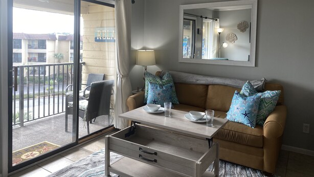 A living room with a sofa, a coffee table, a lamp, and a view of a balcony. - Beautiful Ocean View/ Steps to Beach and Pool (Hilton Head Island)