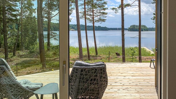 Una terraza de madera con mobiliario de mimbre que da a un lago rodeado de árboles altos.