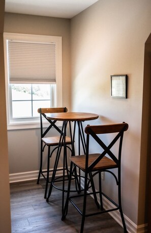 A corner with two wooden tables and chairs, a window with blinds, and a framed picture on the wall. - Studio near Kettle Moraine Forest-hikers paradise! (Kewaskum)