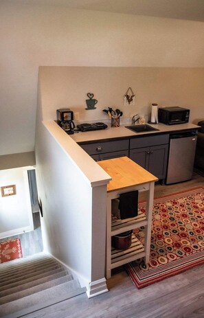 A compact kitchen with a microwave, coffee maker, and a small sink. There's a wooden table and a patterned rug.