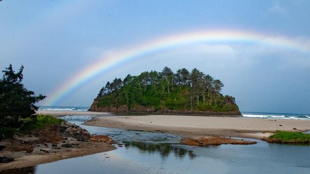Una playa con una pequeña isla cubierta de árboles, un mar tranquilo y un arcoíris en el cielo.