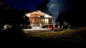 Una cabaña de dos pisos con una escalera destacada que lleva a la entrada, un área de estar exterior con sillas y una chimenea visible que emite humo.